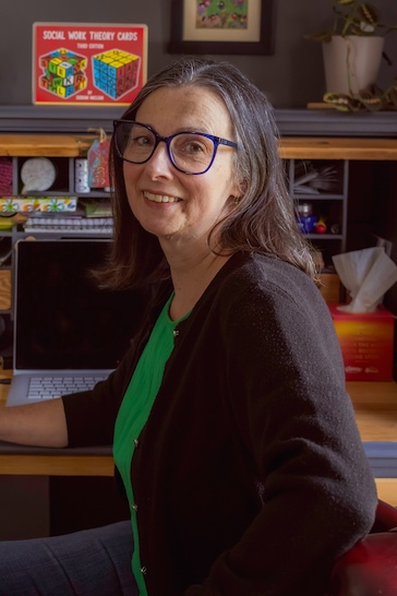 Siobhan Maclean sits at a desk, wearing blue glasses, a green top and cardigan. The desk has a laptop and a number of notebooks, stationary, and her own social work publications.